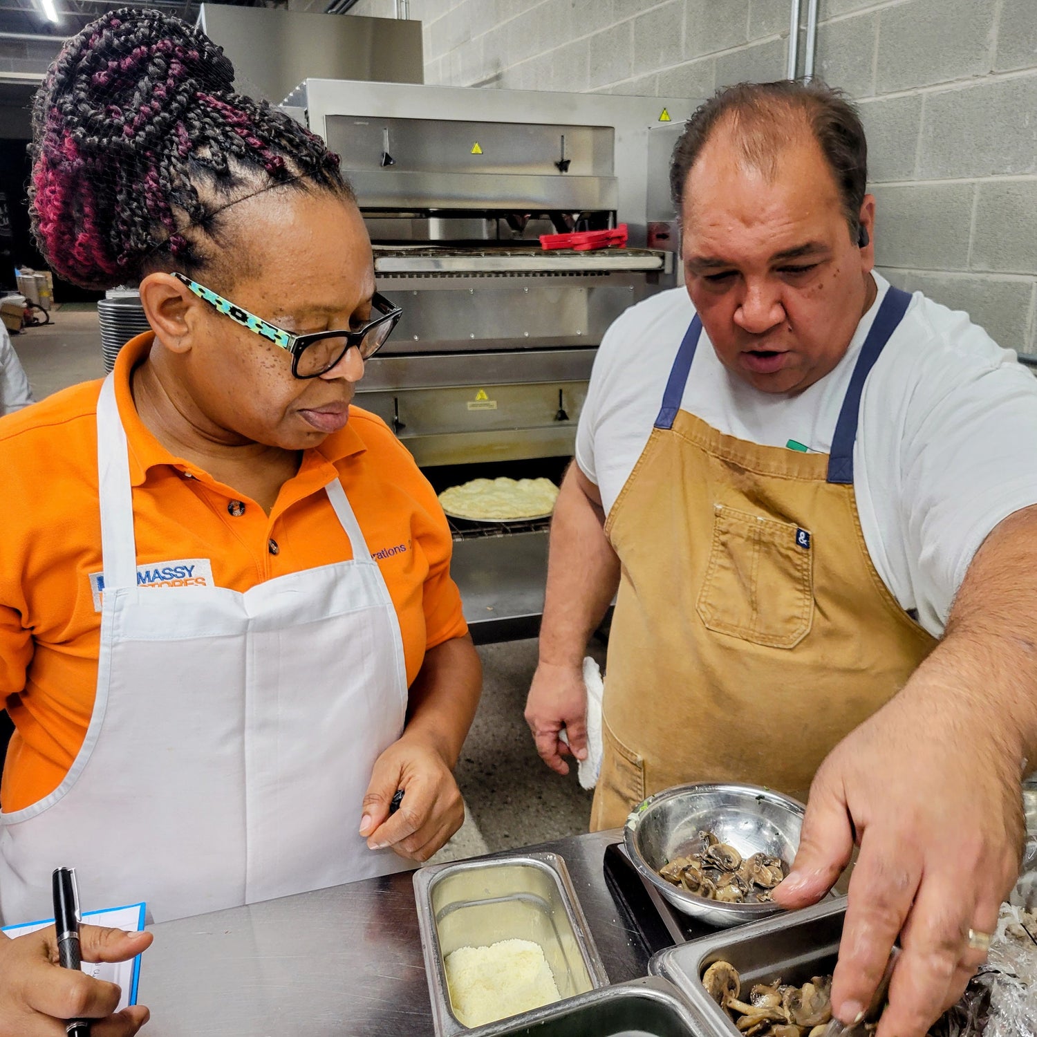 Two people in a kitchen preparing food, one wearing an orange shirt and white apron, the other in a white shirt and brown apron.