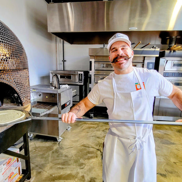 Man in a kitchen with a pizza oven, smiling at the camera.